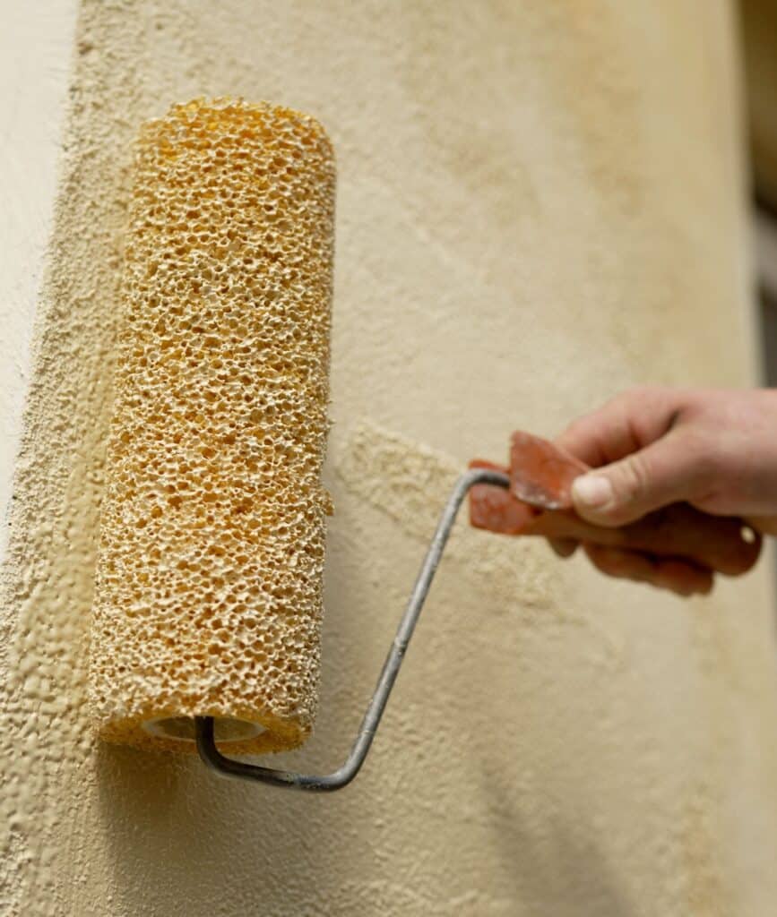Application Peinture Mur Texturé avec Rouleau Mousse Main appliquant une couche de peinture texturée beige clair sur un mur avec un rouleau en mousse à grosses alvéoles.