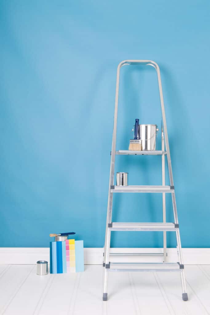 Peinture Murale : Échelle, Couleurs et Matériel Escabeau en aluminium contre un mur bleu clair, avec pots de peinture et nuancier de couleurs sur le sol blanc.